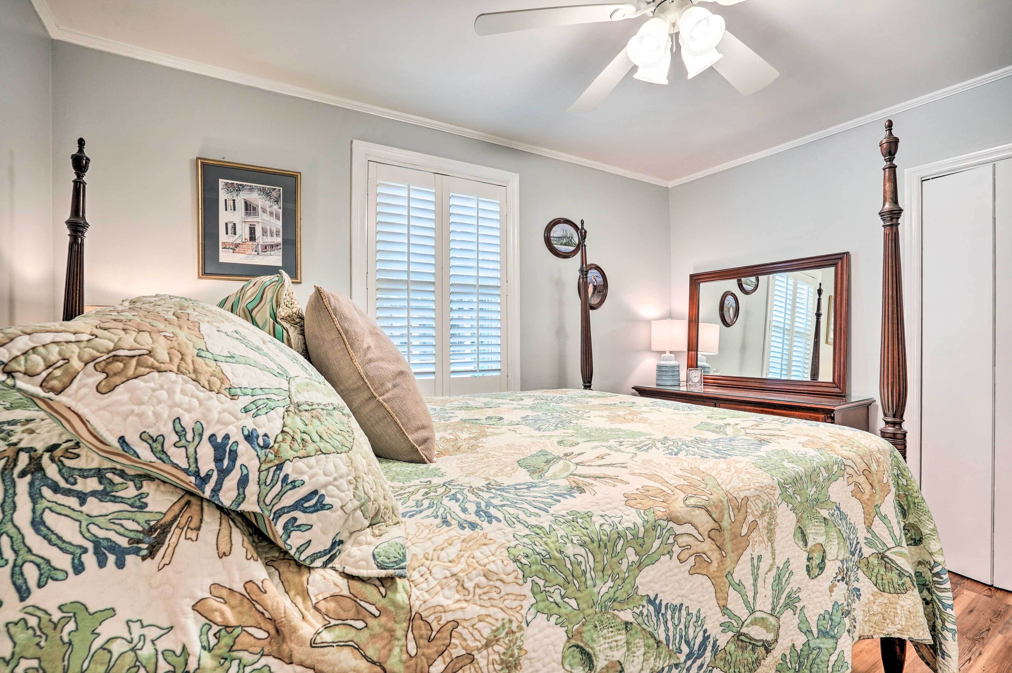 A bedroom with a bed, a ceiling fan, a window with blinds, a bedside table with a lamp, and a mirror.