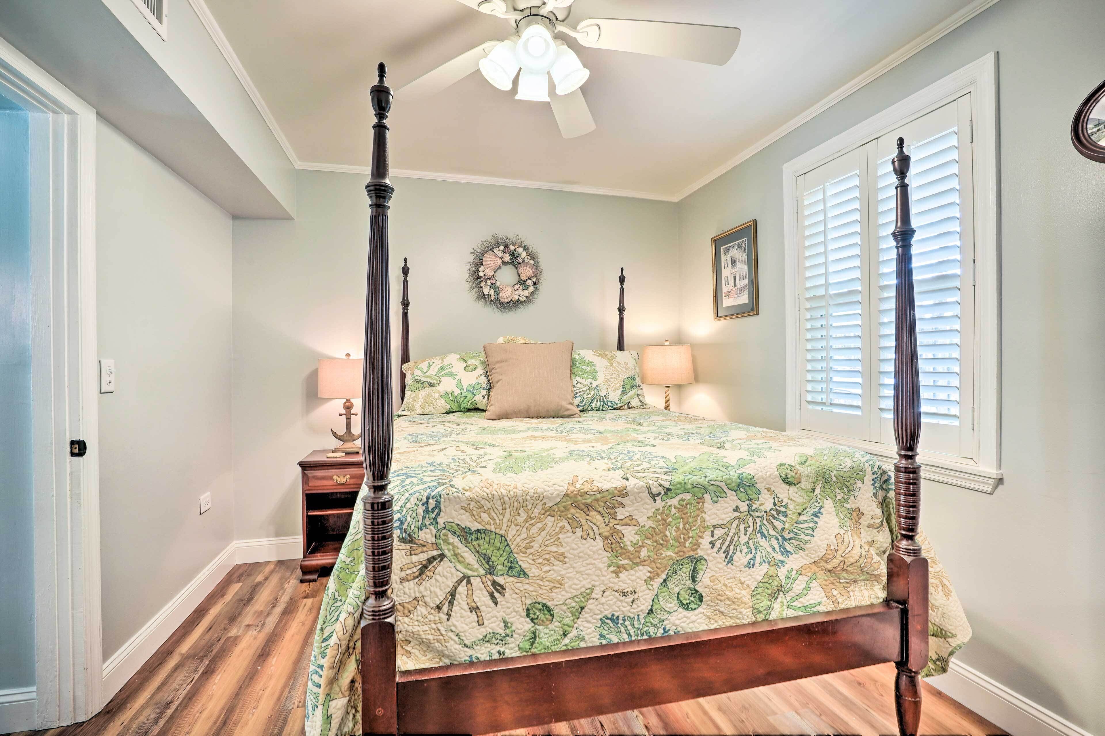 A bedroom with a four-poster bed, a ceiling fan, a nightstand with a lamp, and a window with blinds.