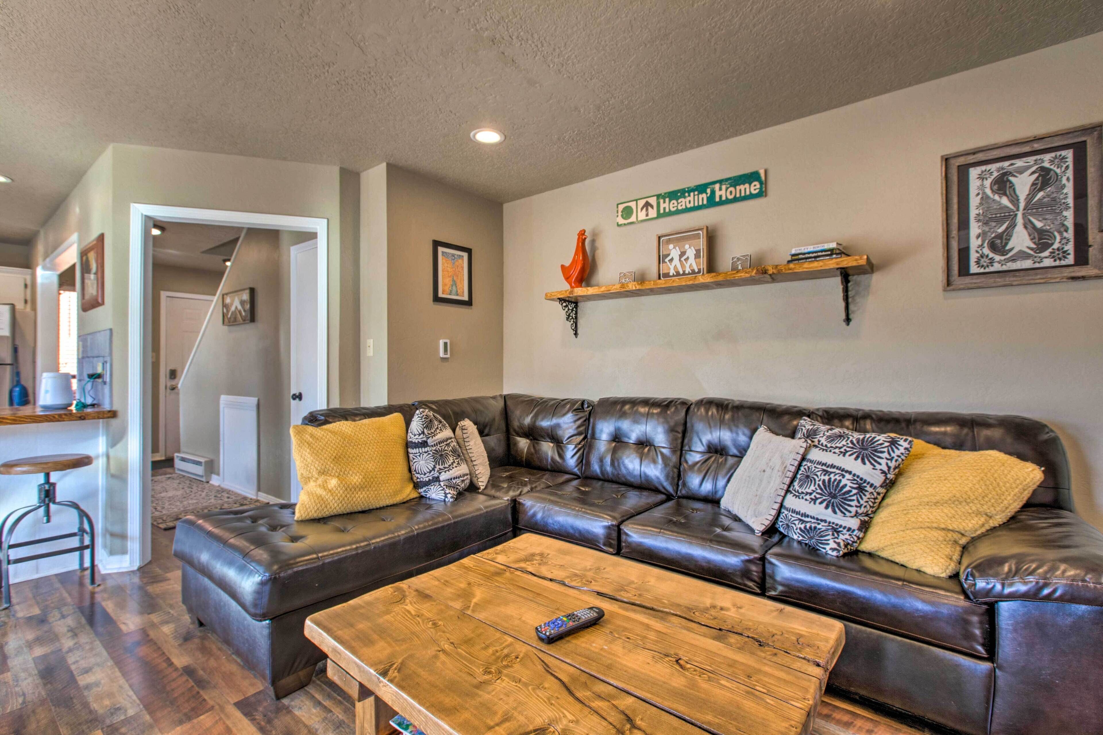 A living room with a dark leather sectional sofa, a wooden coffee table, and a kitchen area in the background.