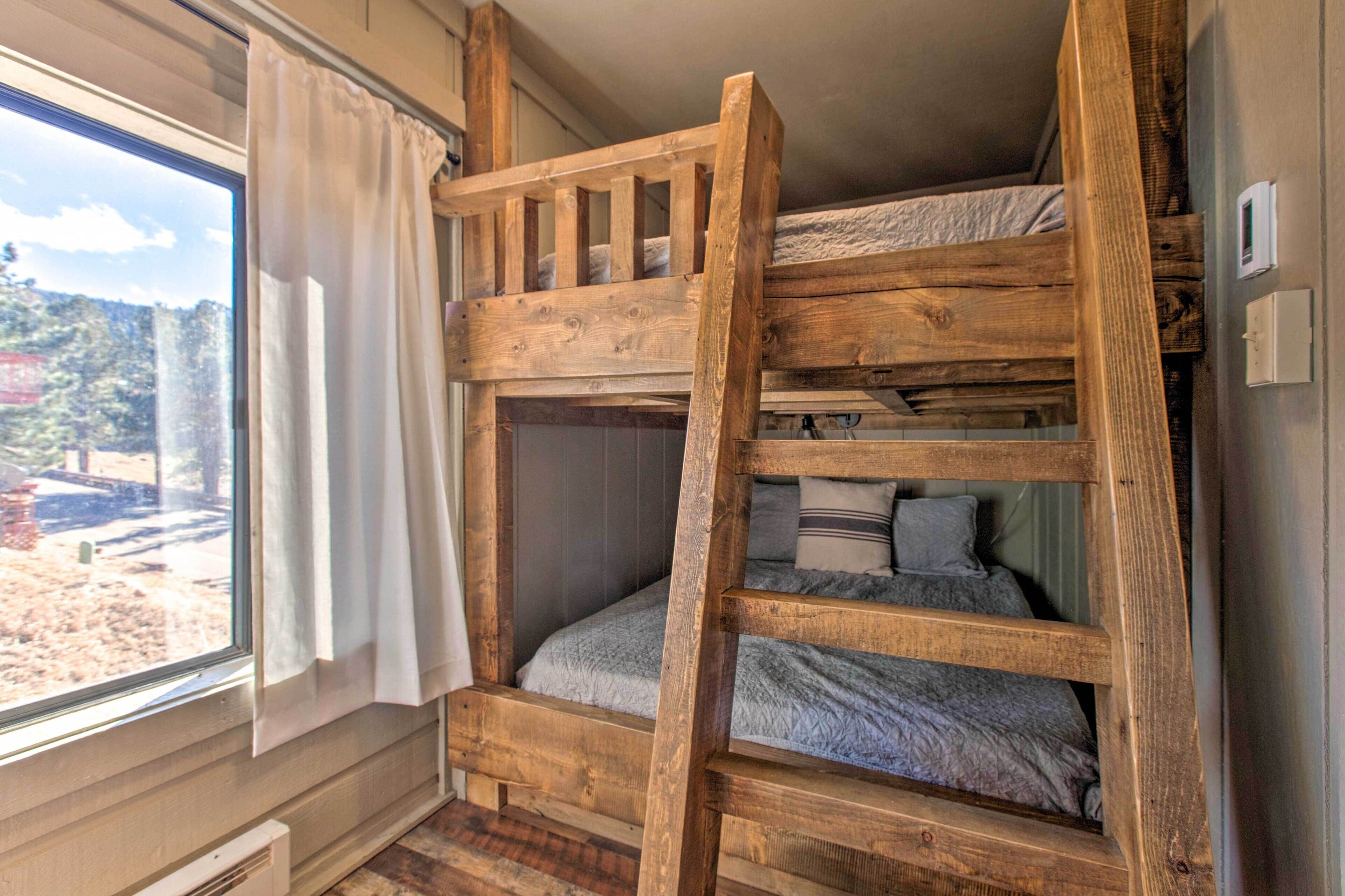 A bunk bed with a wooden ladder, a window with curtains, and a view of a forested area.