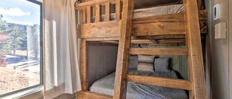 A bunk bed with a wooden ladder, a window with curtains, and a view of a forested area.