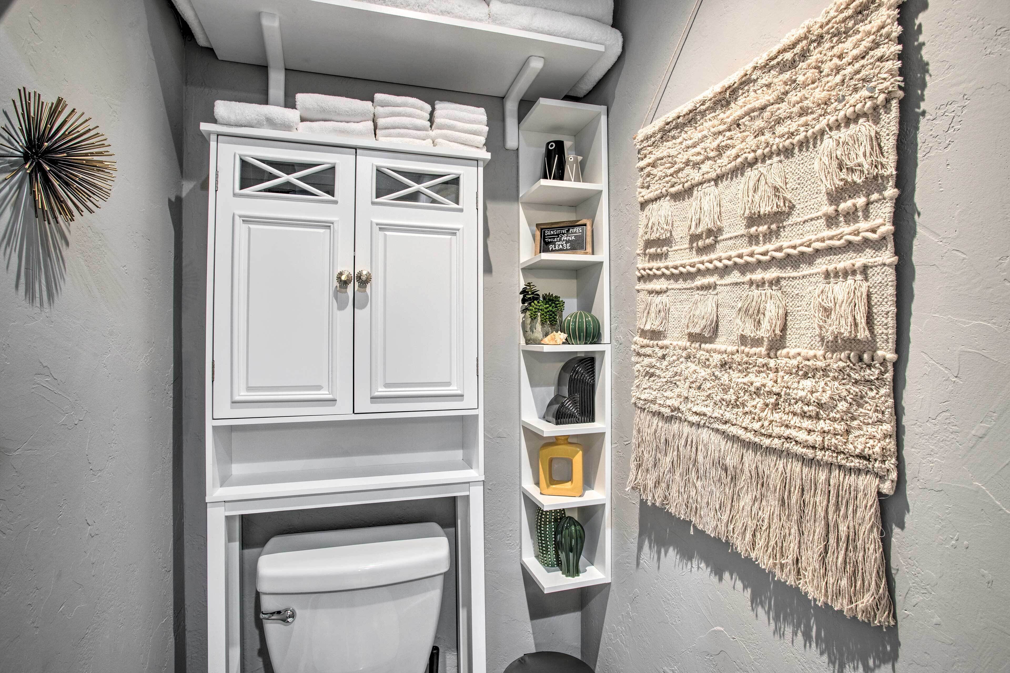 A compact bathroom with a white cabinet, shelves with toiletries, and a textured wall hanging.