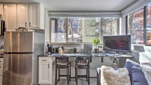 A modern kitchen with white cabinets, a stainless steel refrigerator, a marble countertop, two bar stools, and a large window with a view of trees and buildings. - Telluride Studio in Downtown - Walk to Gondola! (Telluride)