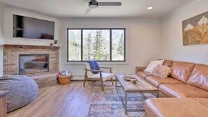 A modern living room with a fireplace, leather sofa, and a large window with a view of trees. - Frisco Mountain Hub: Walk to Main Street (Frisco)