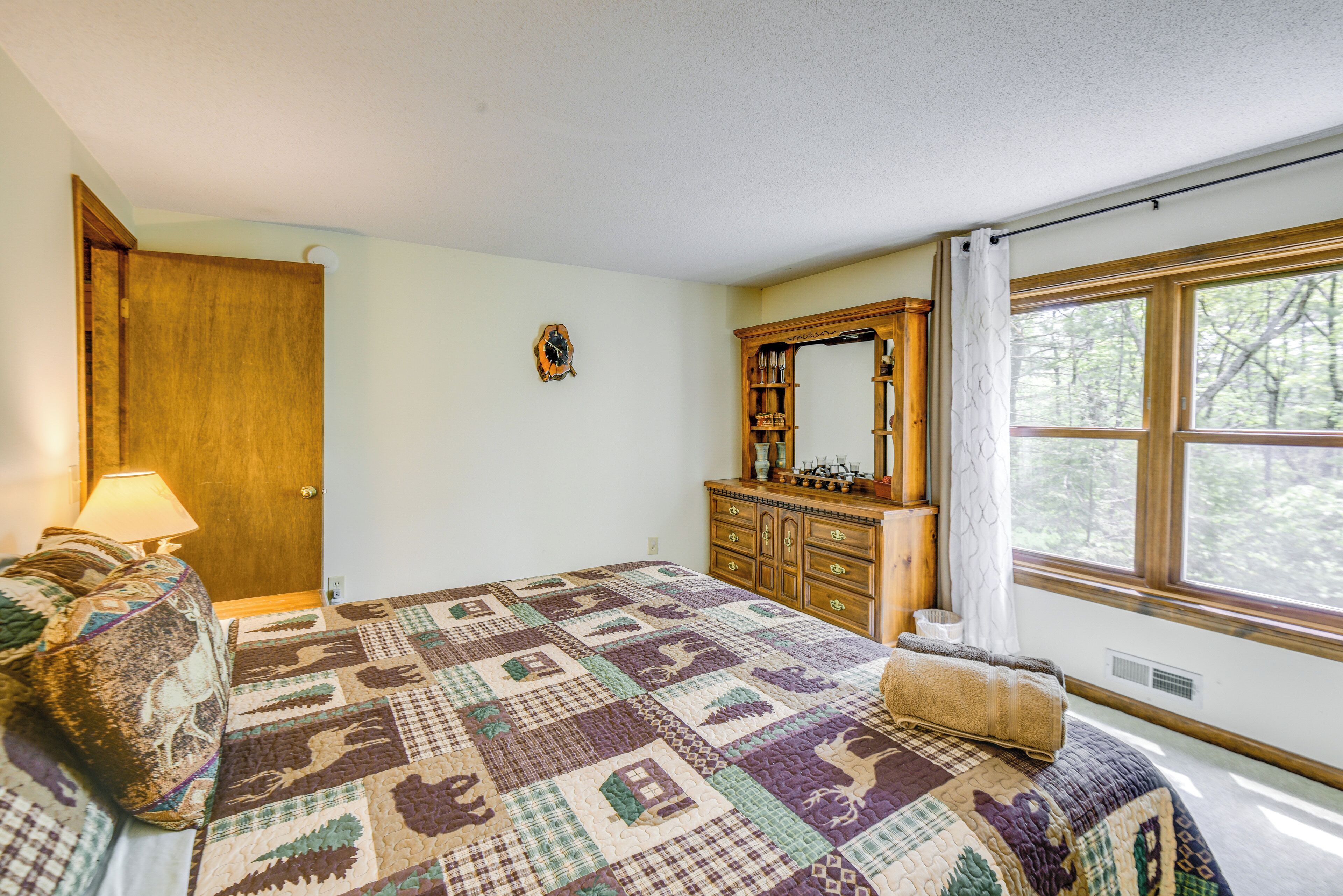 Une chambre à coucher avec un grand lit, une commode, un miroir et une fenêtre donnant sur les arbres.