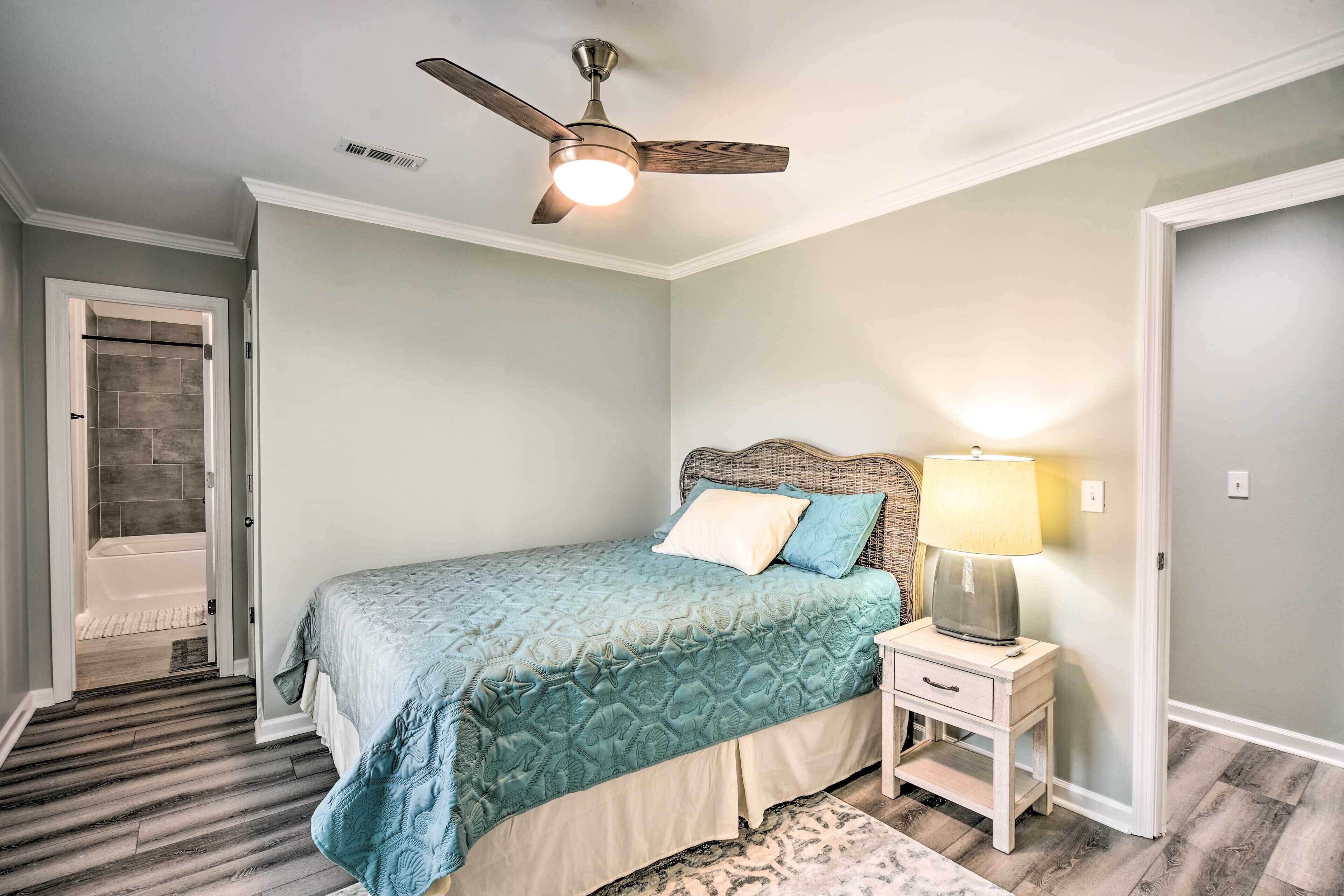 A bedroom with a bed, a nightstand, a lamp, a ceiling fan, and a door leading to a bathroom.
