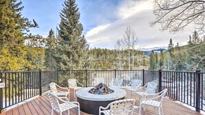 A deck with a circular fire pit and white chairs, surrounded by a black railing, overlooking a forested area with mountains in the distance. - Cozy Ski-in/out Winter Park Studio w/ Hot Tubs! (Winter Park)