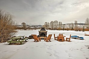 A snow-covered outdoor area with a stone fireplace, wooden chairs, and a picnic table. - Fish, Hike & Ski: Riverfront Granby Cabin! (Granby)