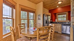 A wooden dining table with chairs, a kitchen with a refrigerator and dishwasher, and large windows with blinds. - Fish, Hike & Ski: Riverfront Granby Cabin! (Granby)