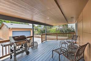 Um deck coberto ao ar livre com churrasqueira, cadeiras de vime e vista para uma casa ao fundo.