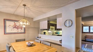 Een eetgedeelte met een houten tafel, stoelen, een kroonluchter, een ingelijst schilderij, en een keuken met kasten en apparatuur.