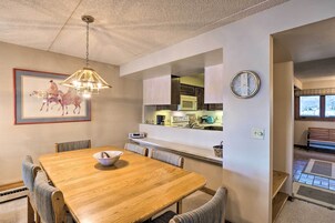 Un comedor con una mesa de madera, sillas, una araña, un cuadro enmarcado y una zona de cocina con armarios y electrodomésticos.