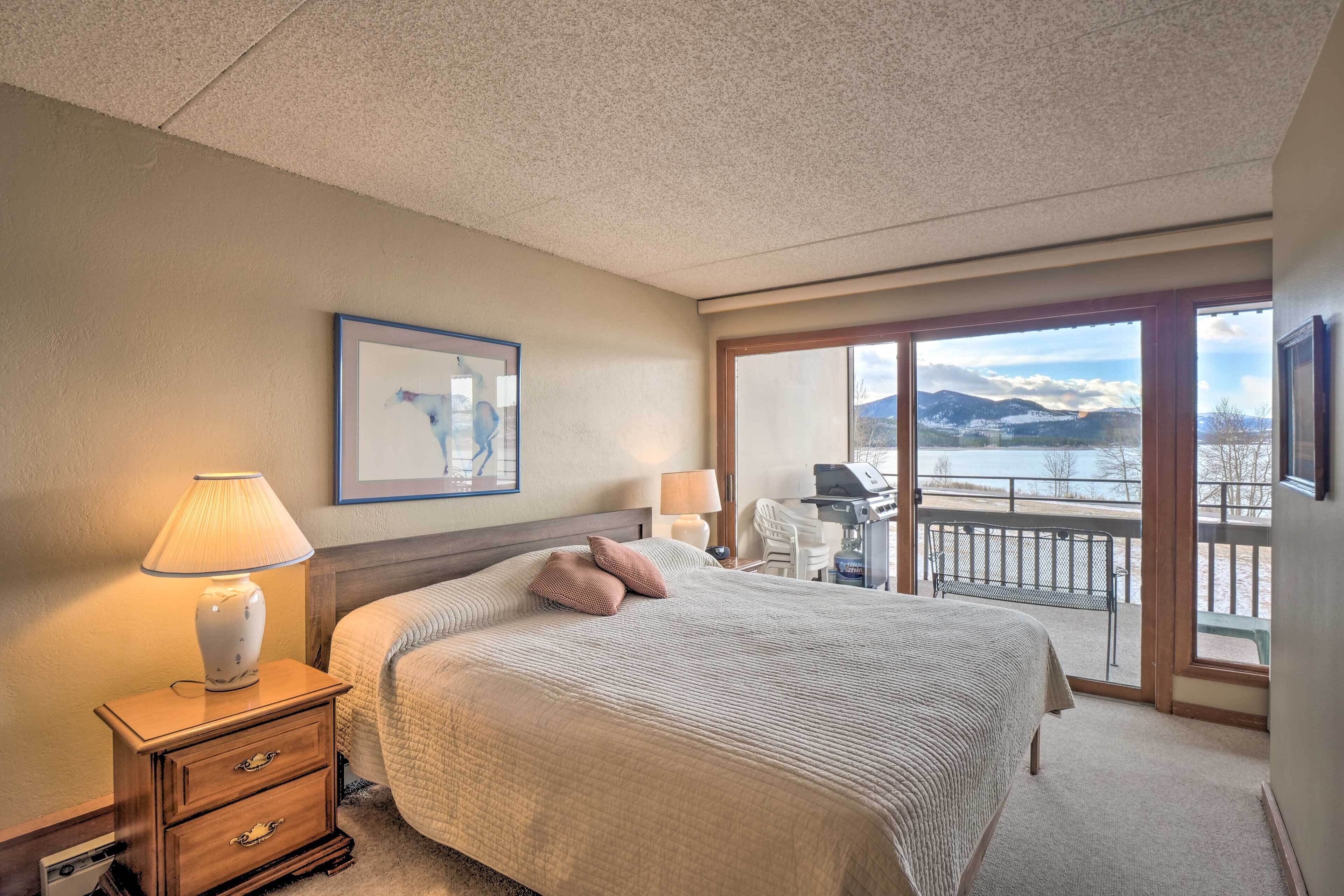 A bedroom with a bed, bedside lamps, a nightstand, a desk, and a view of a balcony with a scenic landscape.