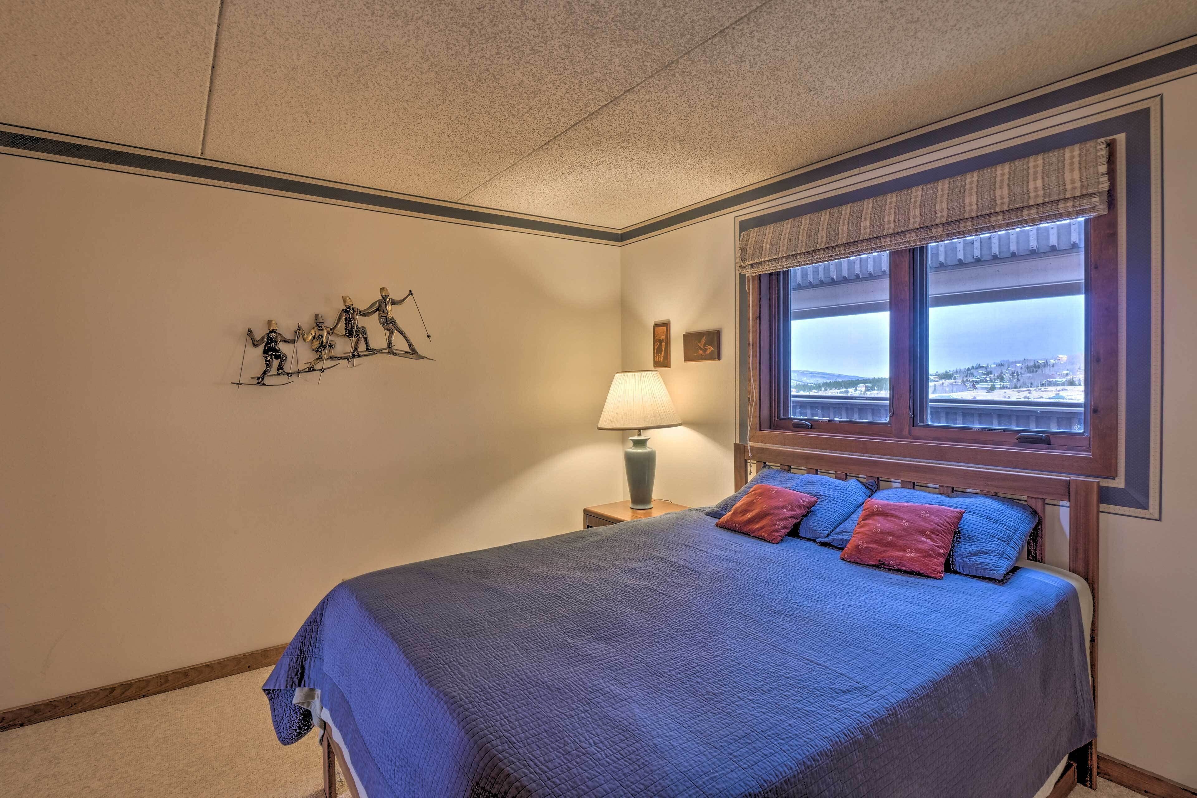 A bedroom with a large bed, a nightstand with a lamp, a window with a view, and a wall decoration.