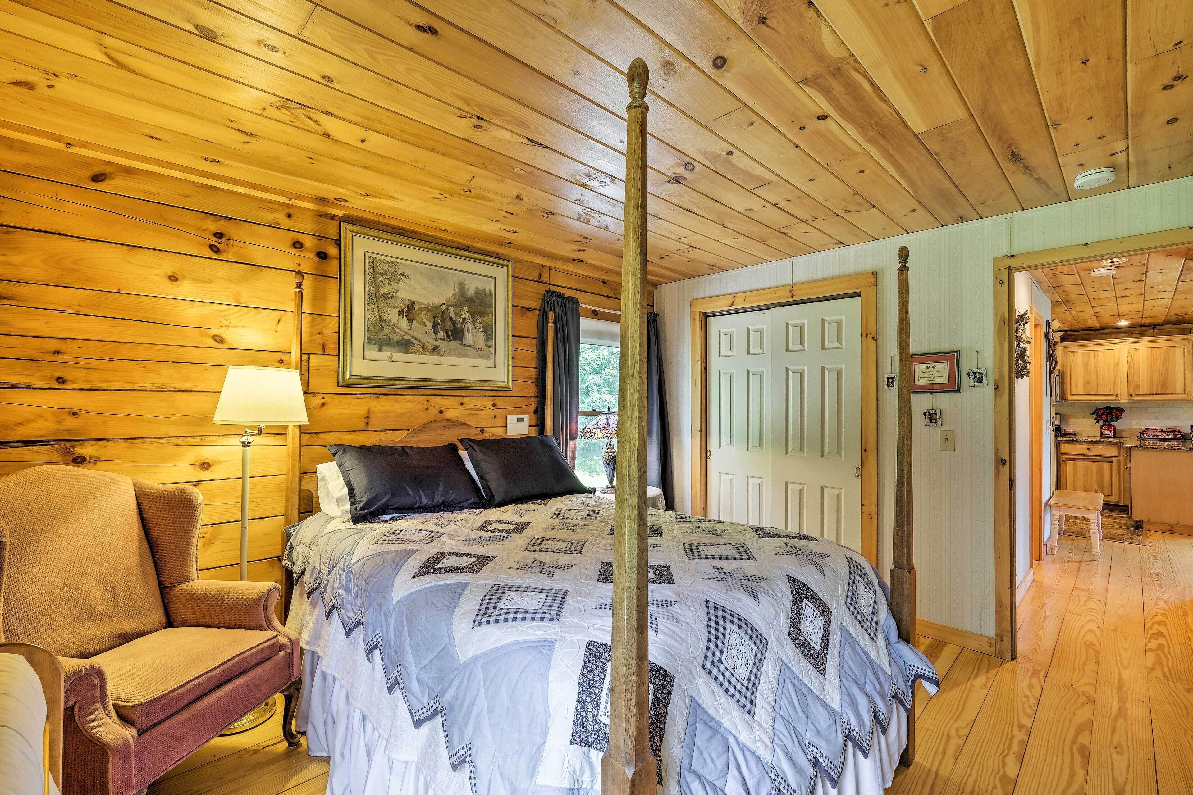 A wooden-paneled room with a bed, a chair, a lamp, and a door leading to a kitchen area.