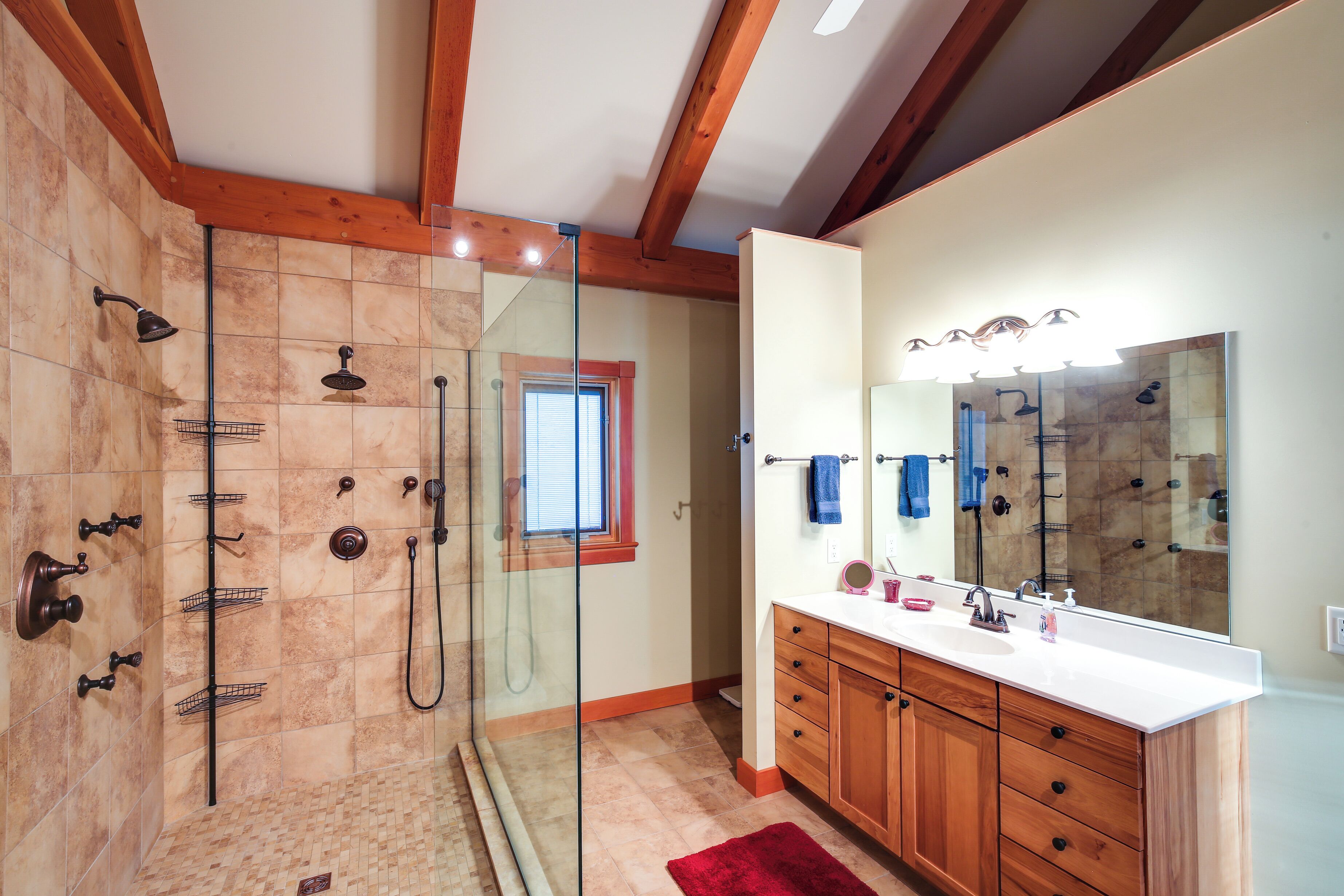 Un baño con ducha envuelta en vidrio, armarios de madera, un espejo grande y un tapete rojo.