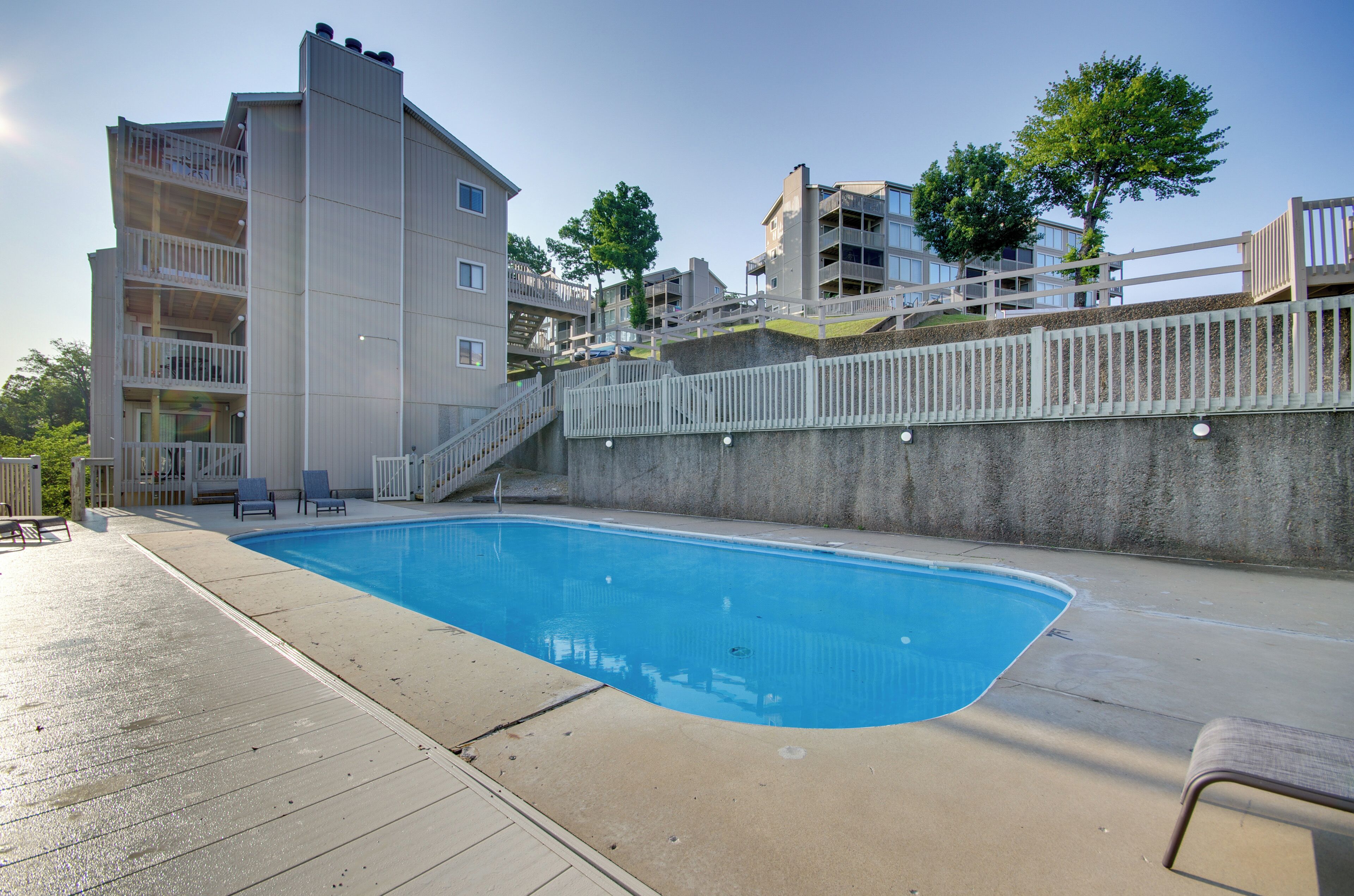 A swimming pool with a concrete deck, a white fence, and a multi-story building in the background.