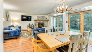 A cozy living room with a fireplace, a dining table, and a large window offering a view of the outdoors. - Renovated Ski-in/ski-out Peru Condo at Bromley Mtn (Peru)
