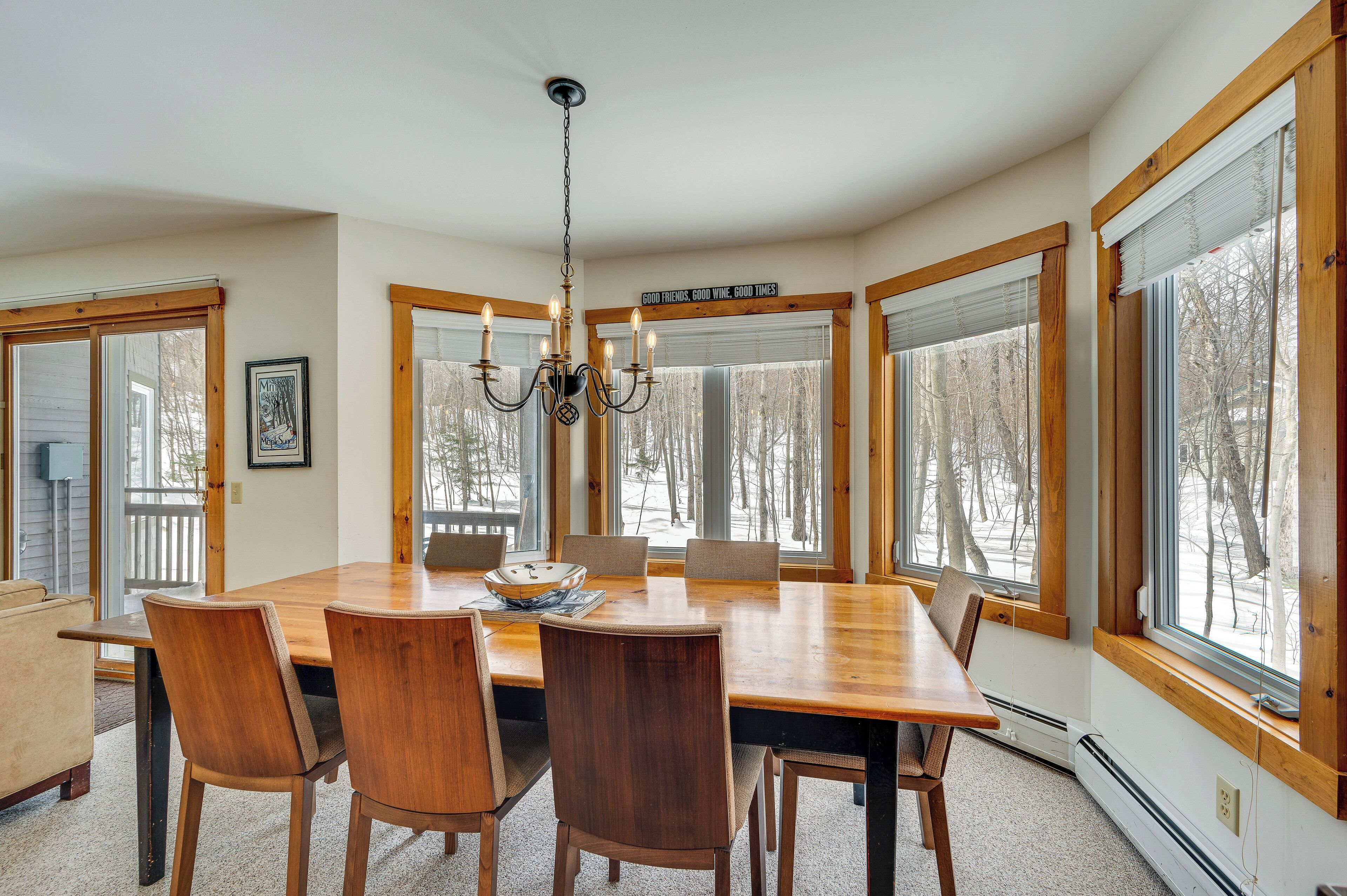 Une salle à manger avec une grande table en bois, six chaises, un lustre et de grandes fenêtres à cadres en bois.