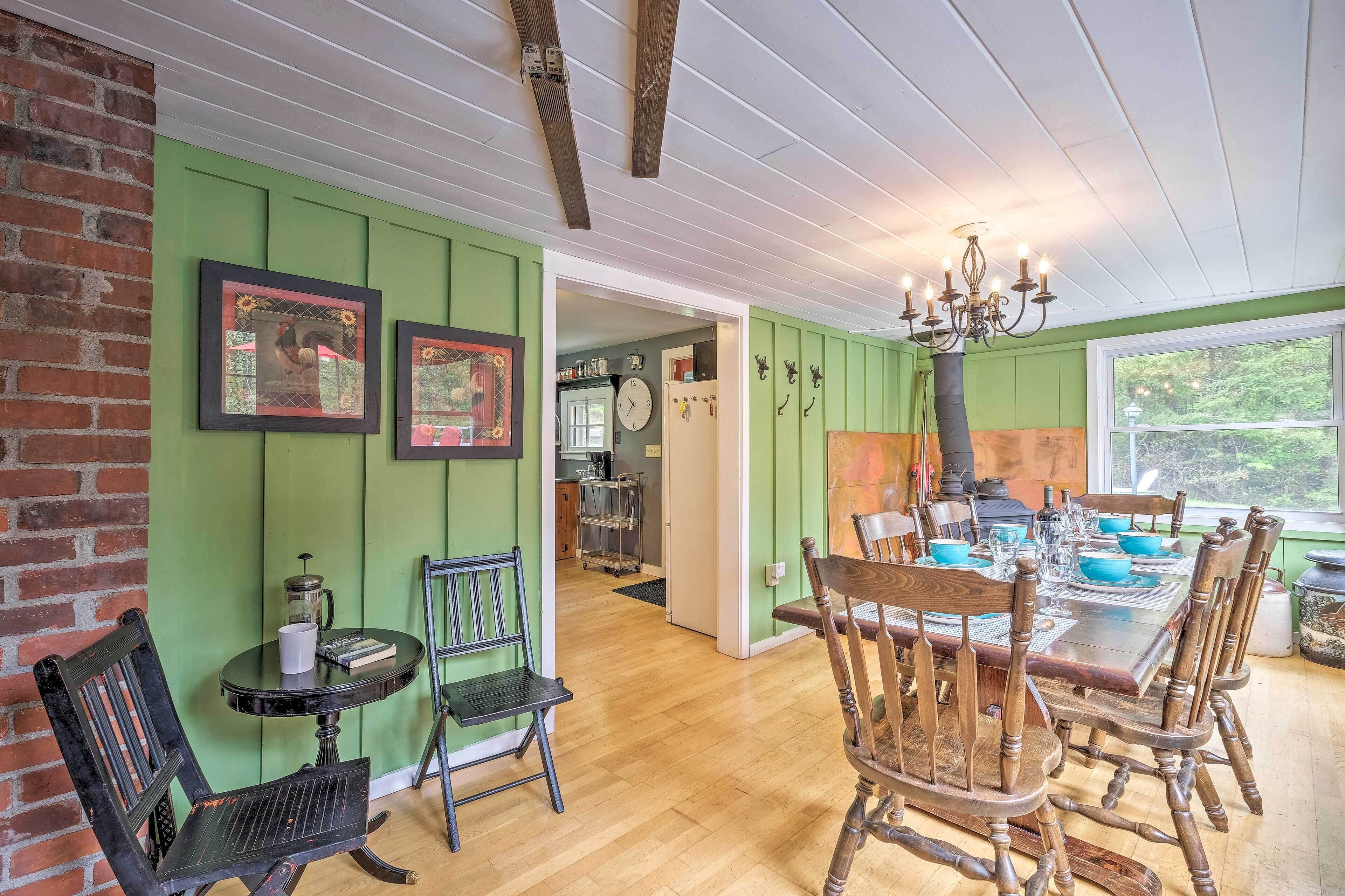 A dining area with a wooden table set for six, surrounded by wooden chairs. A chandelier hangs above the table, and the walls are painted green.