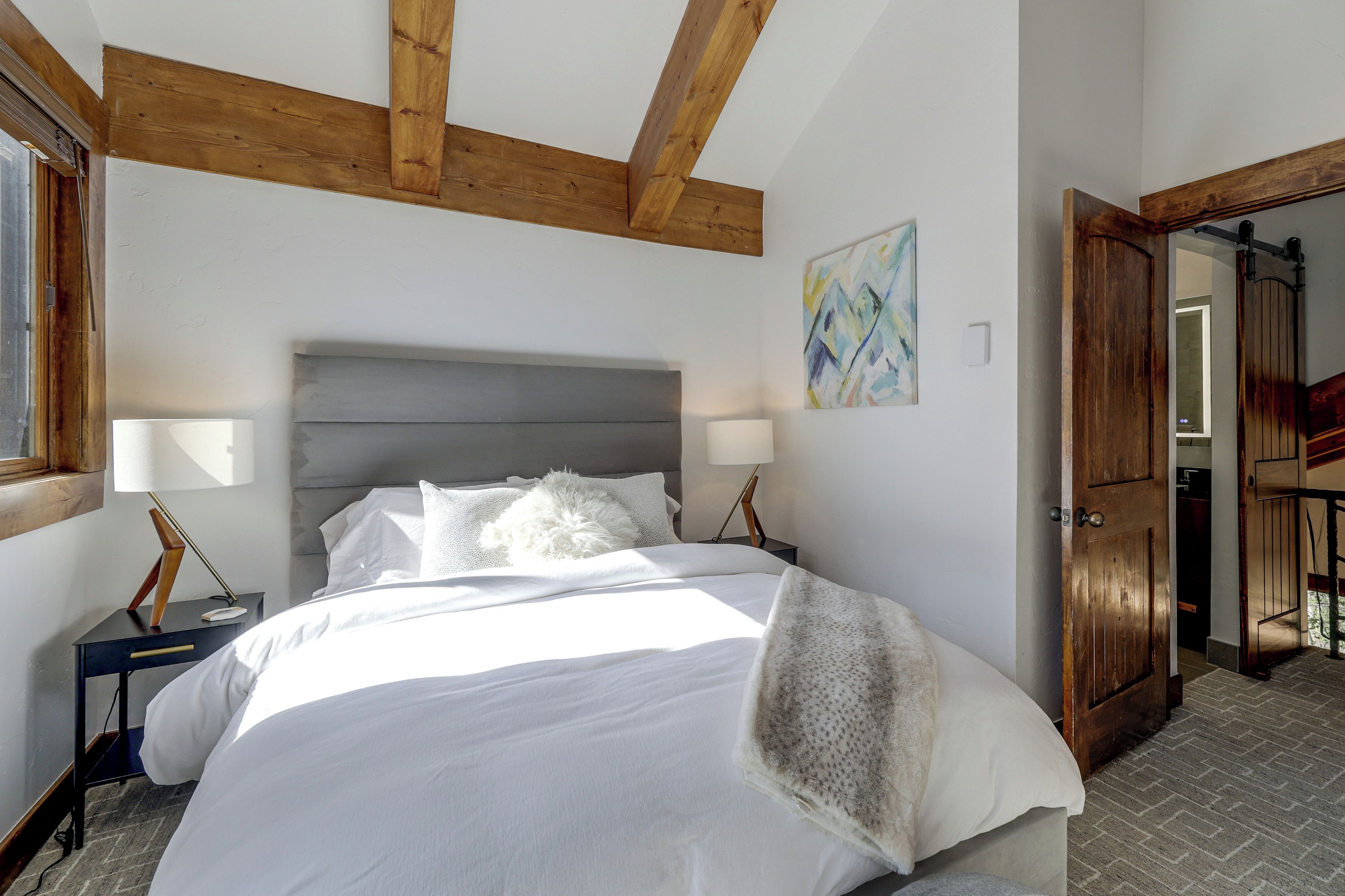 A bedroom with a large bed, wooden beams, a nightstand, a lamp, and a door leading to another room.