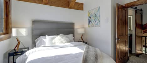 Ein Schlafzimmer mit einem großen Bett, Holzbalken, einem Nachttisch, einer Lampe und einer Tür zu einem anderen Raum.