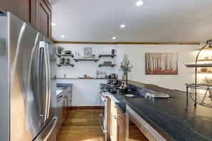 A modern kitchen with a stainless steel refrigerator, wooden cabinets, and a black countertop. There are built-in shelves with various kitchen items and a painting of a forest on the wall. - Newly Renovated Vail Home - Walk to Booth Falls Tr (Vail)