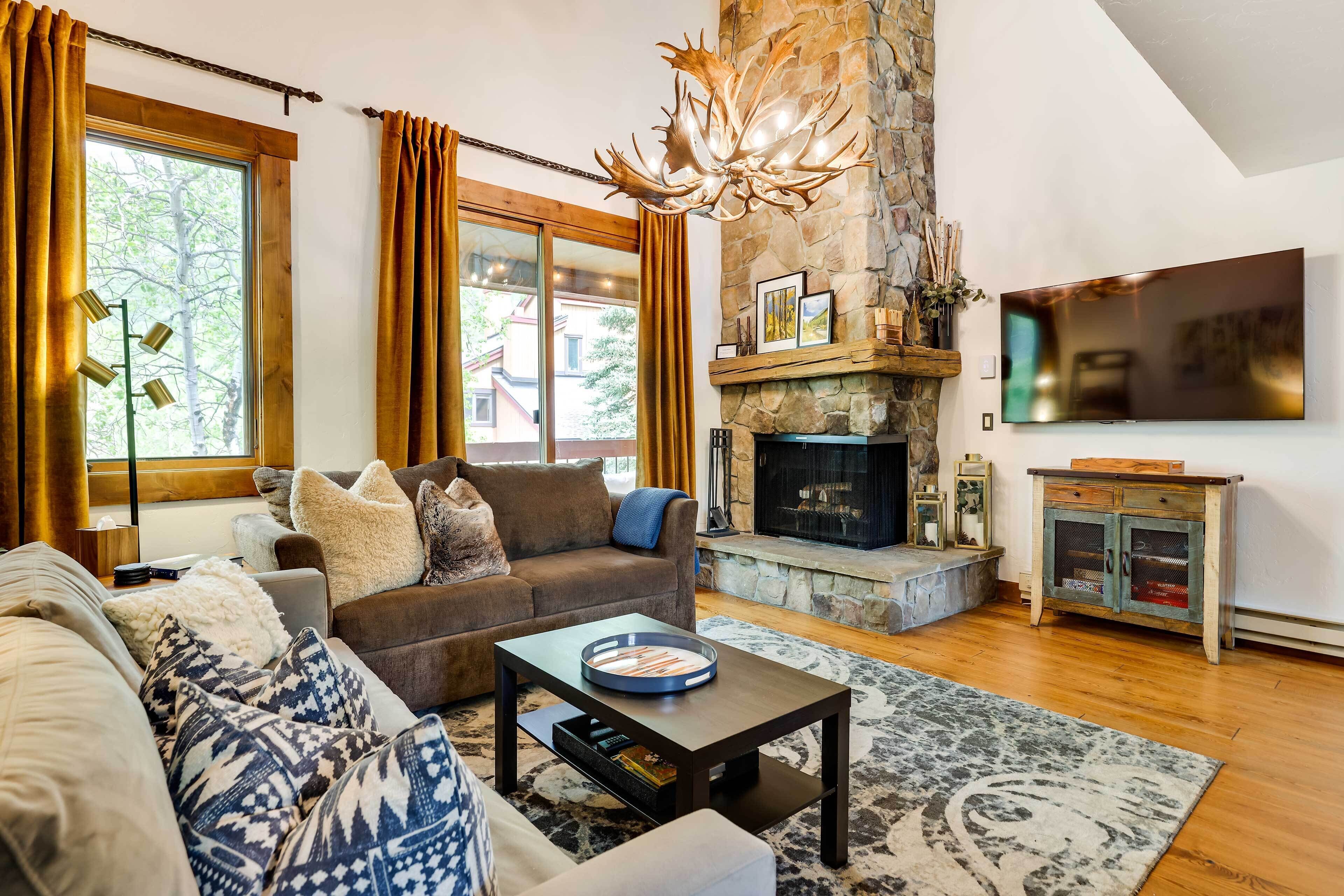 A living room with a stone fireplace, a flat-screen TV, and a large window with curtains.