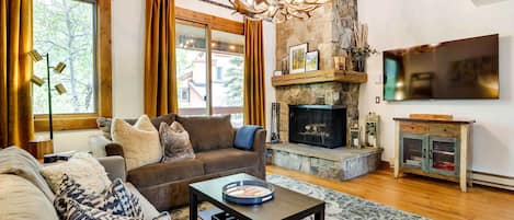A living room with a stone fireplace, a flat-screen TV, and a large window with curtains.