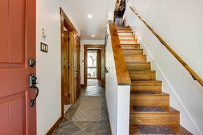 Un escalier en bois avec une rampe, une porte rouge avec une poignée noire et un tapis à motifs sur le sol.