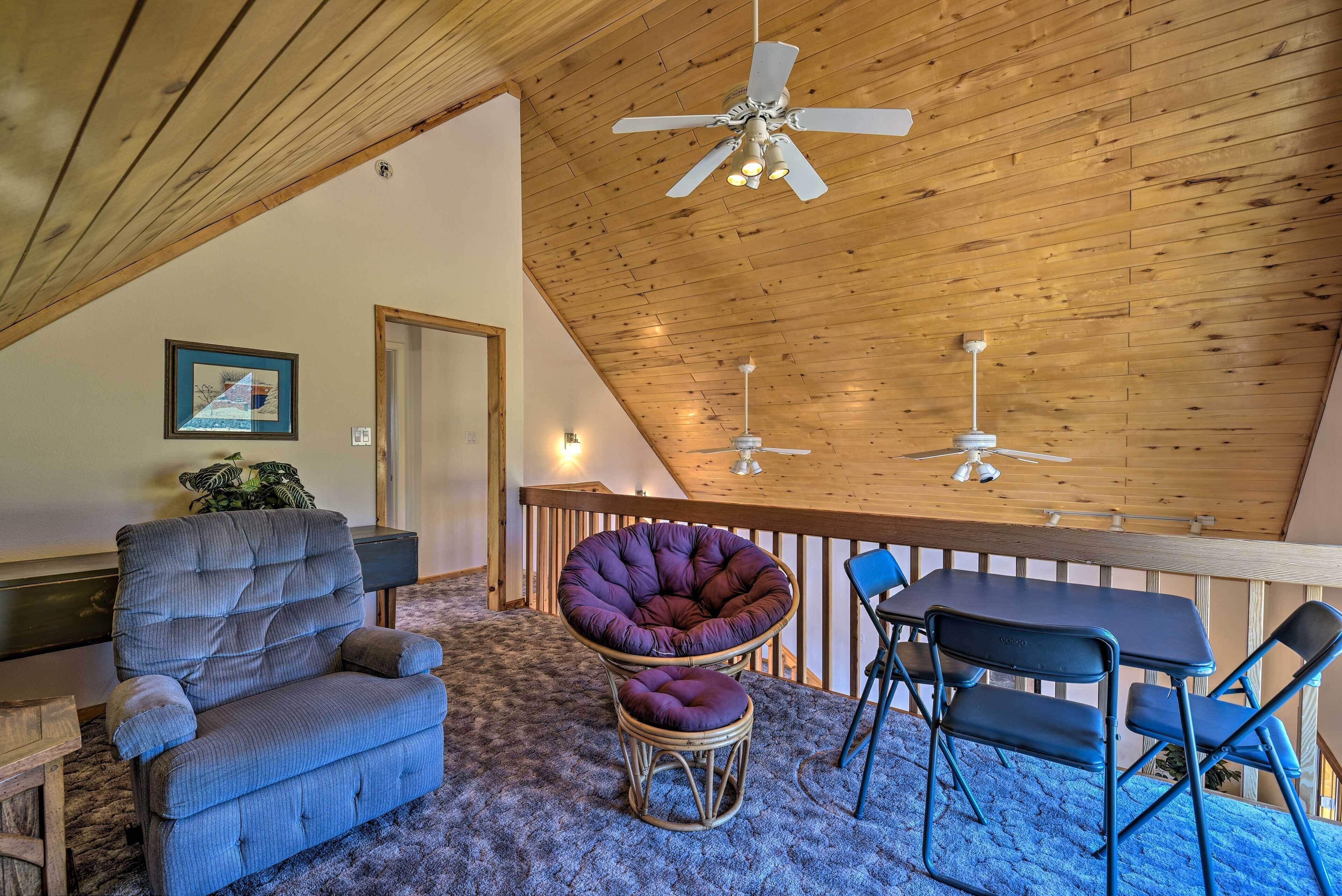 A cozy loft bedroom with a wooden ceiling, a ceiling fan, a dining table with chairs, a cushioned chair, and a small round table.