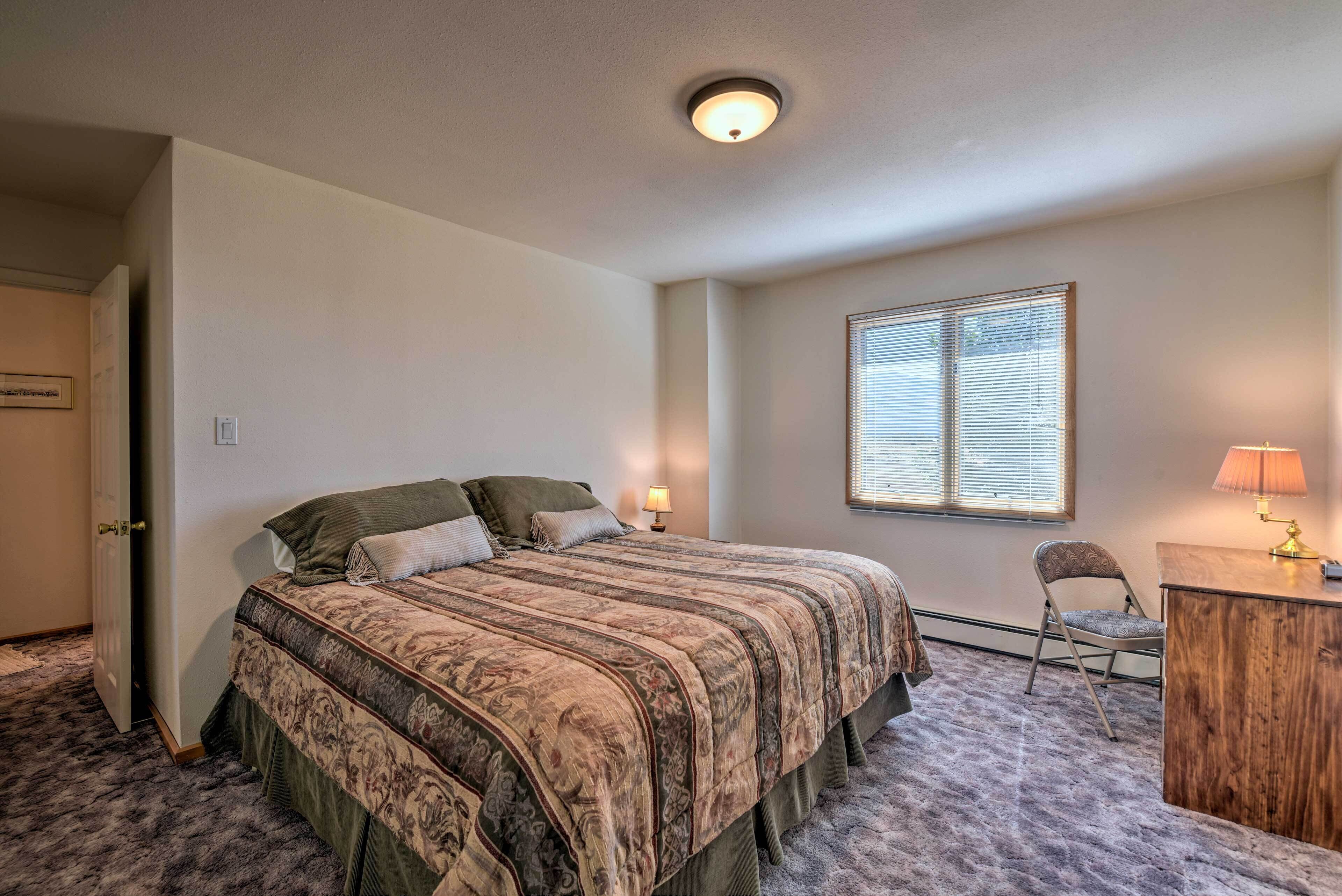 A bedroom with a bed, a desk, a chair, a window with blinds, and a door.