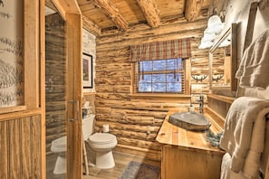 A rustic cabin bathroom with wooden walls, a wooden vanity, a window with plaid curtains, and a mirror with a towel hanging on the right.