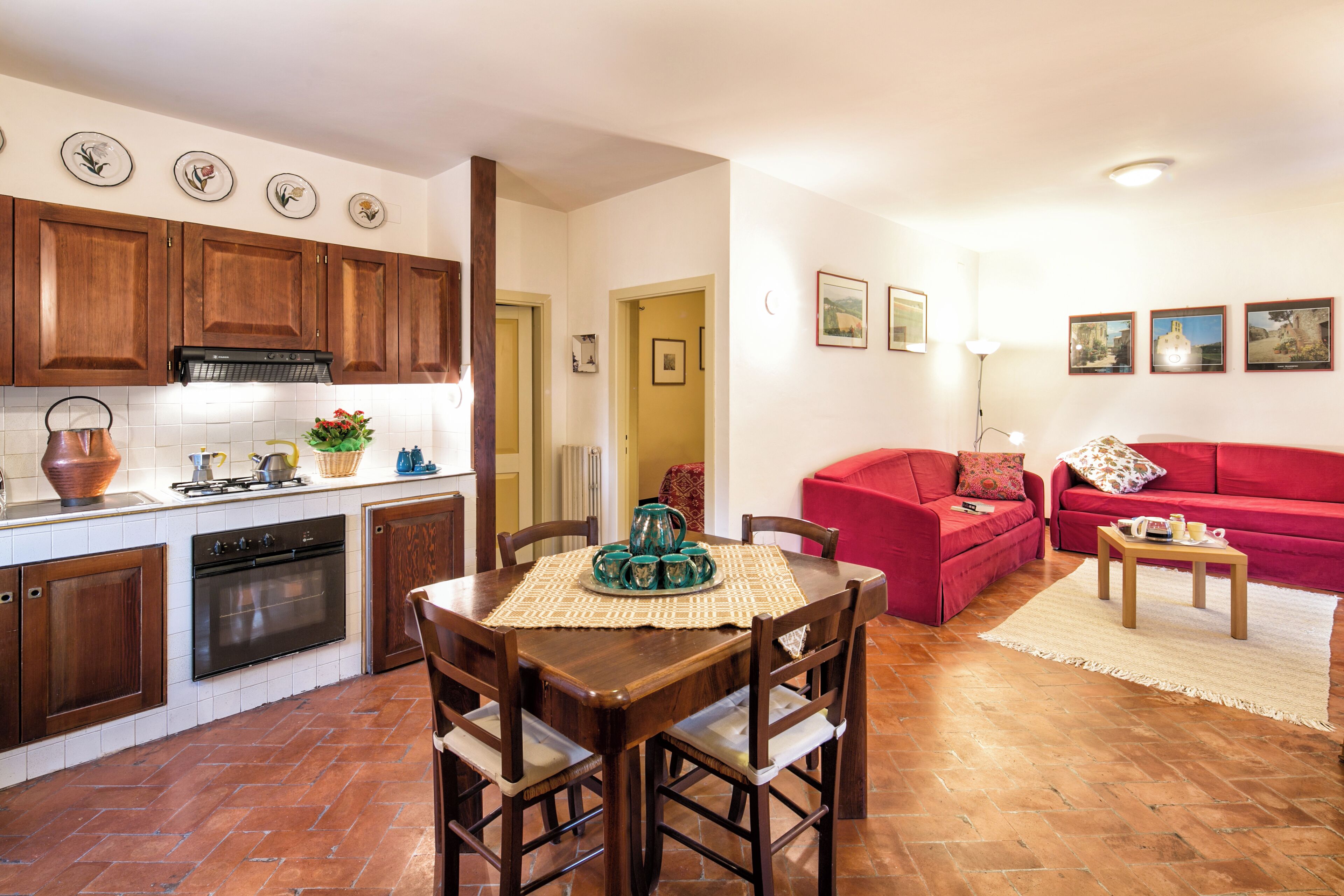 A compact kitchen with wooden cabinets and a dining area with a table and chairs. There is a red sofa in the living area.