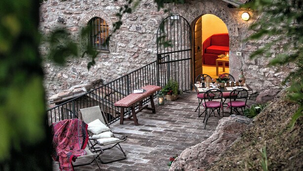A stone building with a staircase leading to a dining area with a red sofa and a table set for two. - Apartment with terrace (Rasiglia)