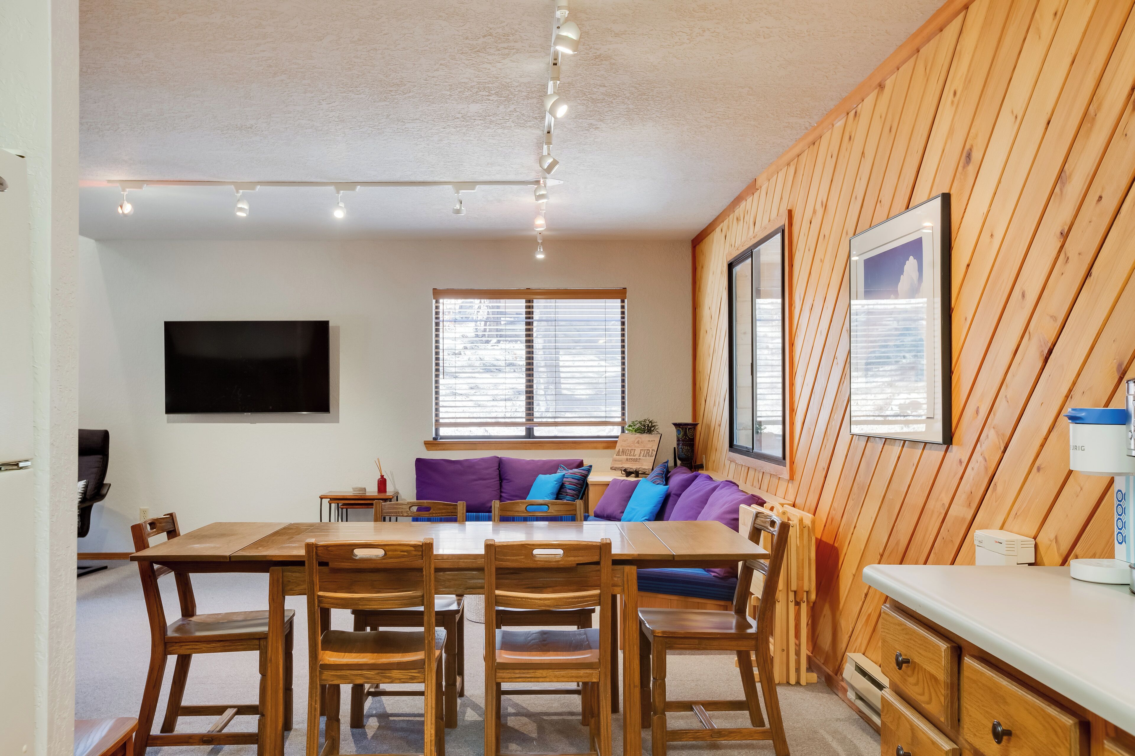 A wooden-paneled room with a dining table, chairs, a sofa with cushions, a mounted TV, and a window with blinds.