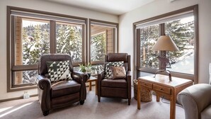 A room with two leather armchairs, a wooden desk, and a lamp. Large windows provide a view of snow-covered trees.