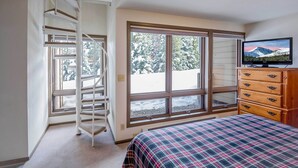 A bedroom with a bed, a dresser, a television, a window with a view of trees, and a spiral staircase.