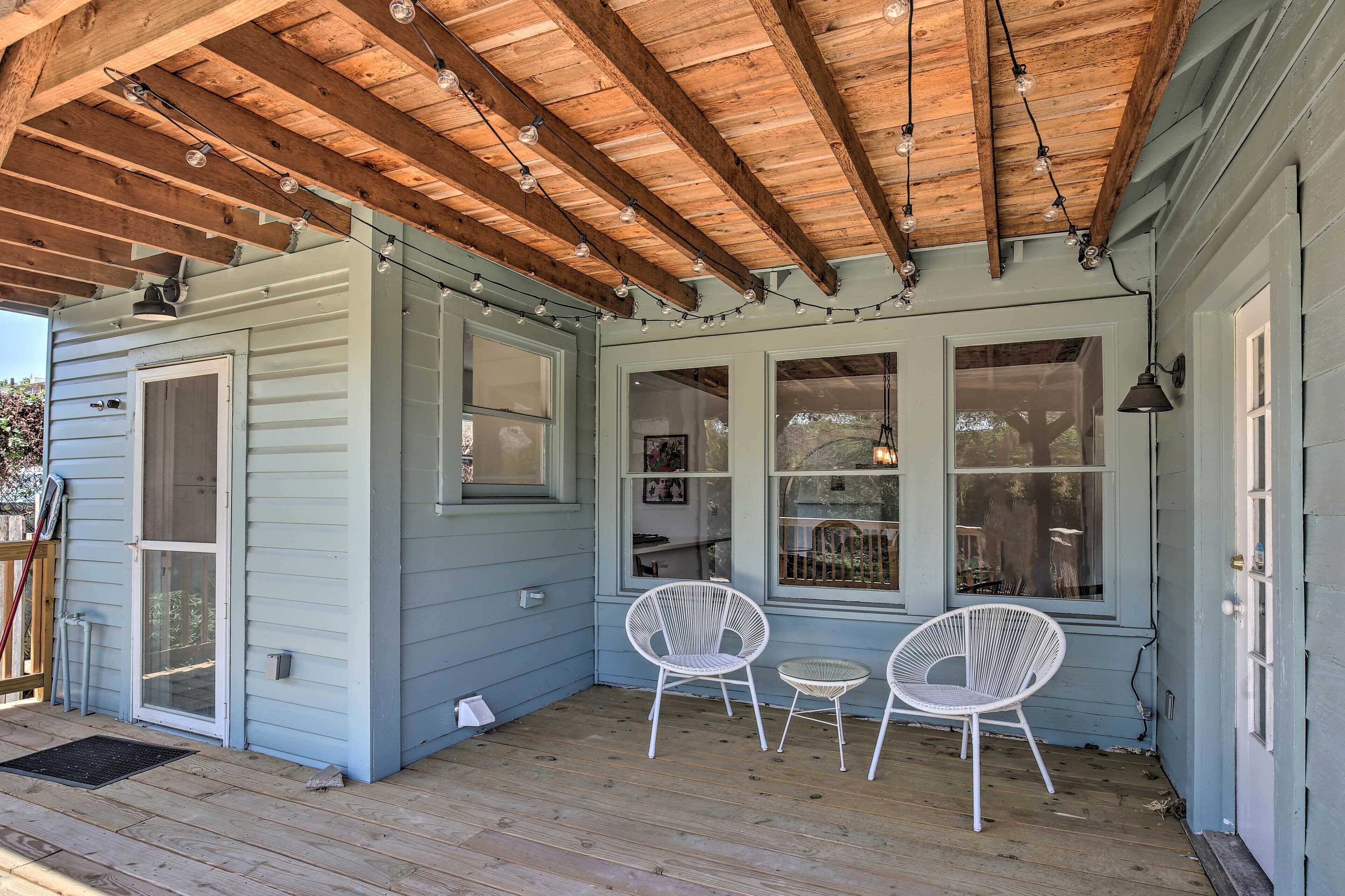Une véranda couverte avec des poutres en bois et des guirlandes lumineuses, deux fauteuils en osier blancs et une petite table.