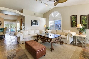 A spacious living room with a large sectional sofa, a coffee table, and a brown ottoman. There are two side tables with lamps, a ceiling fan, and a large window with blinds.