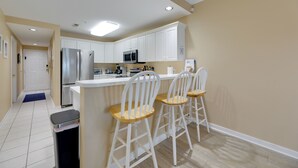 A kitchen with white cabinets, a stainless steel refrigerator, a microwave, and a sink. There are three white bar stools with wooden seats. - Steps to Sand! Family-friendly Ocean City Getaway (Ocean City)