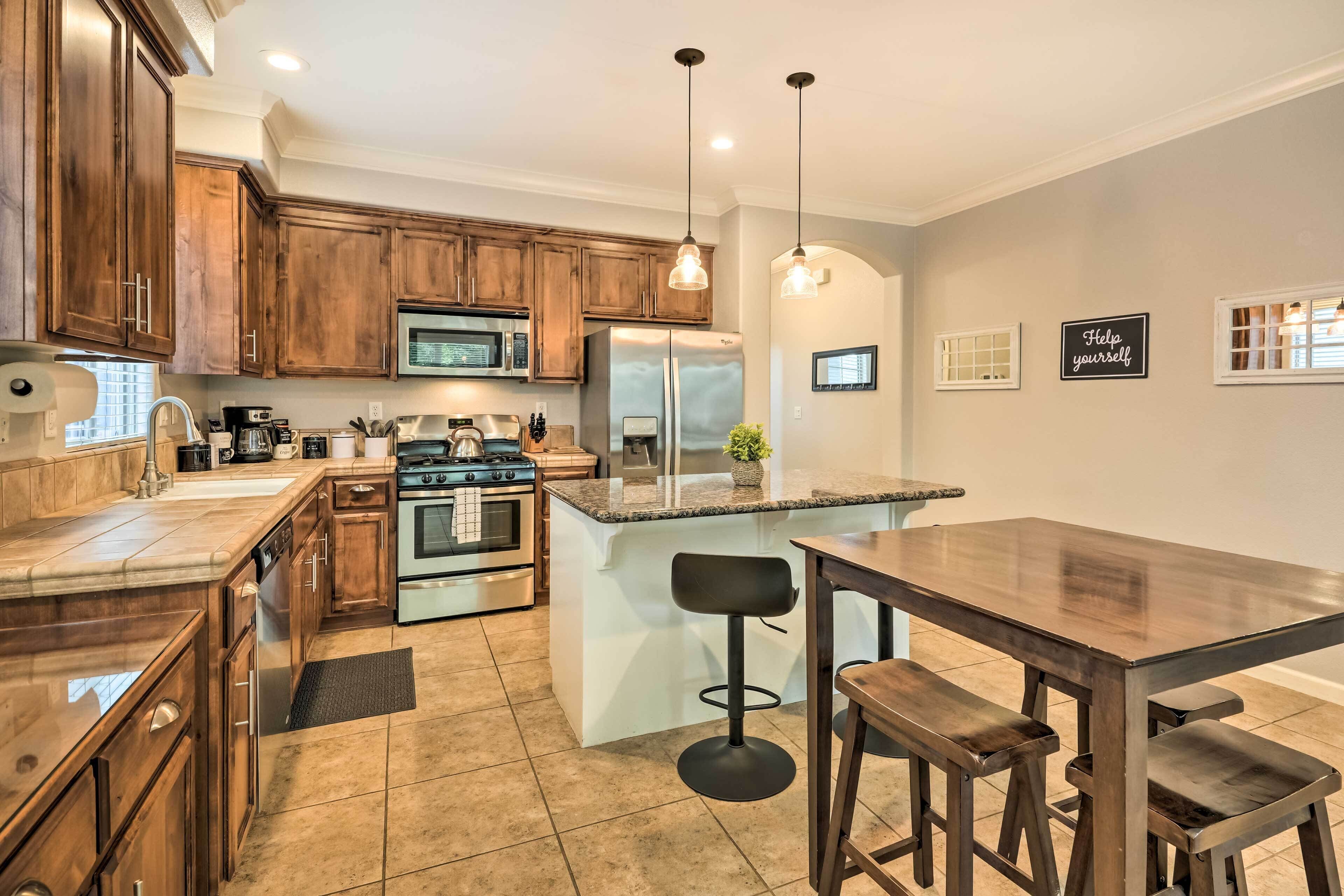 A modern kitchen with wooden cabinets, stainless steel appliances, a central island with bar stools, pendant lighting, and a 'Help yourself' sign on the wall.