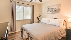 Ein Schlafzimmer mit Bett, Deckenventilator, Fenster mit Vorhängen, Nachttisch mit Lampe und einem an der Wand hängenden Bild.