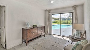A room with a wooden cabinet, a glass table with a lamp, a view of a swimming pool, and a door. - Port Orange Oasis: Close to Beaches & Dining! (Port Orange)