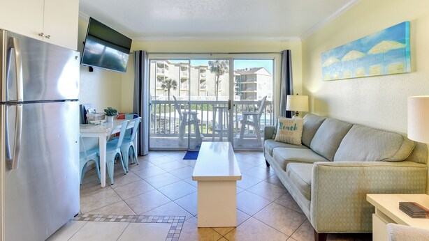 A modern kitchen with a stainless steel refrigerator, a flat-screen TV mounted above the counter, and a dining area with a white table and blue chairs. - Walk to Galveston Beach: Resort Condo w/ Perks! (Galveston)