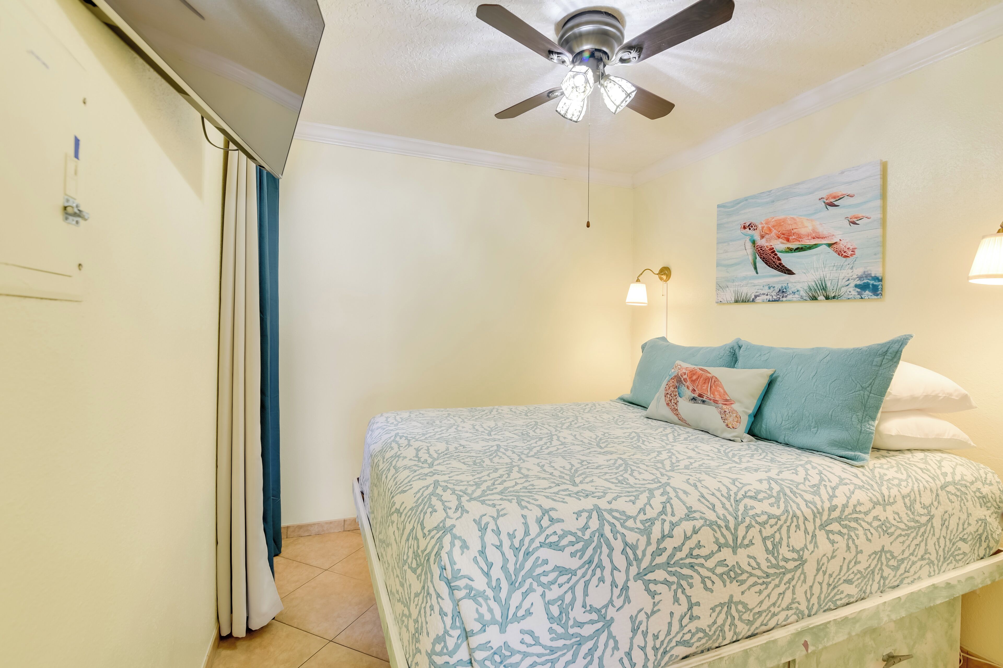A bedroom with a bed, a ceiling fan, a painting of sea turtles, and two wall-mounted lamps.