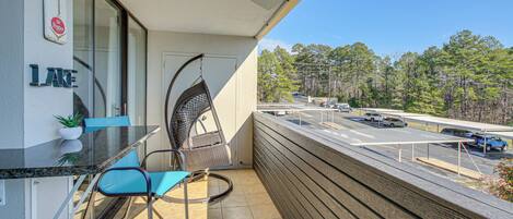 A balcony with a glass table, chairs, a hanging chair, a sign reading "HOT and COLD", and a view of a parking lot and trees.