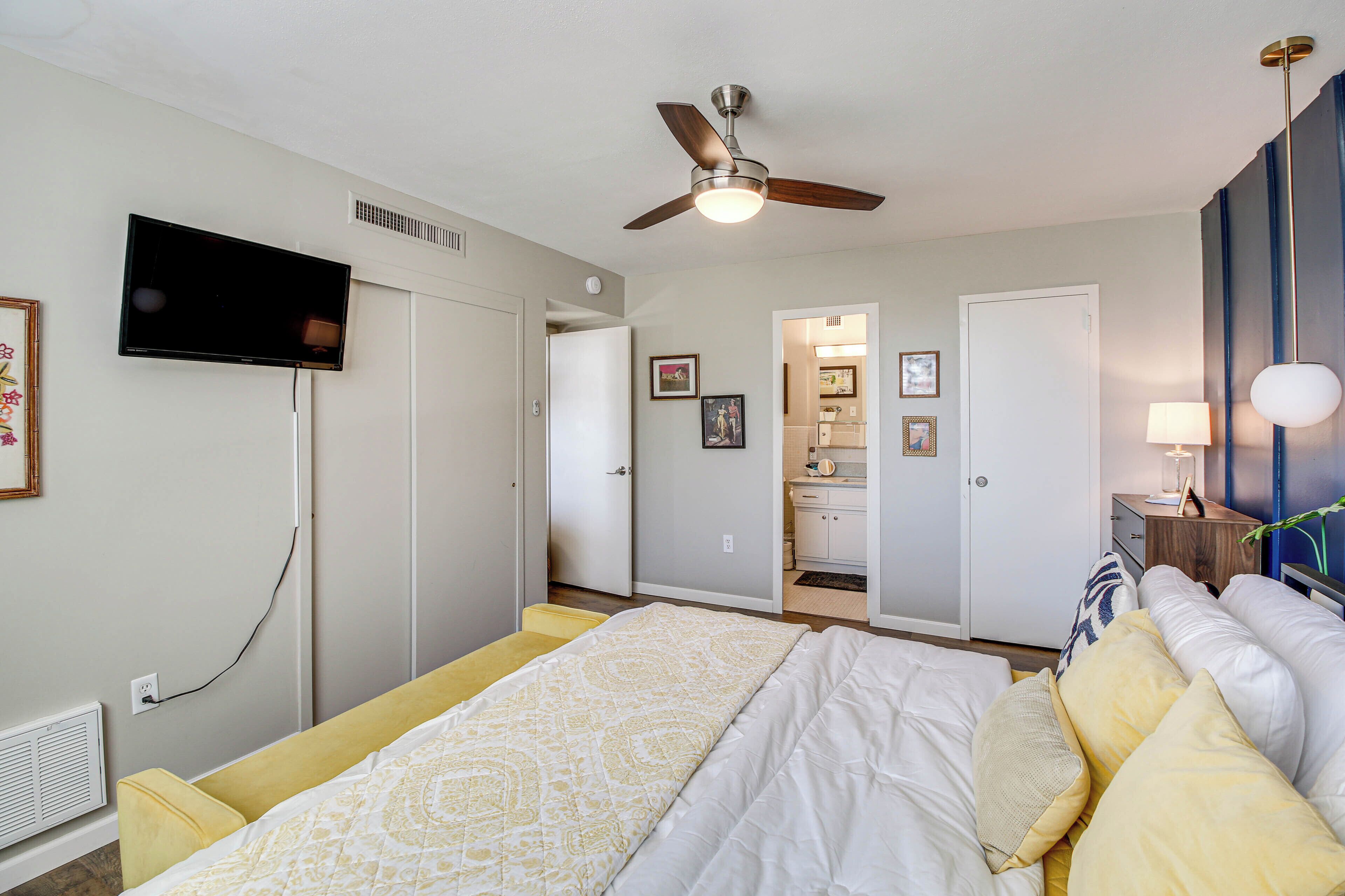 A bedroom with a bed, a ceiling fan, a TV, and a bathroom visible through an open door.