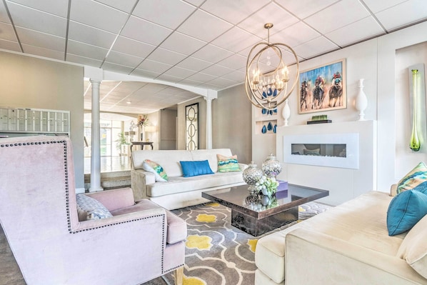 A spacious living room with a white sofa, a pink armchair, and a beige sofa. There is a coffee table with decorative items and a chandelier.