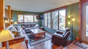 A cozy living room with a sofa, armchair, and coffee table, large windows, and a flat-screen TV.